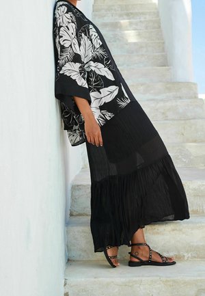 Zwarte kimono met witte bladprint, gedragen over een zwarte geplooide maxirok. Gecombineerd met zwarte sandalen met gespen en studs.