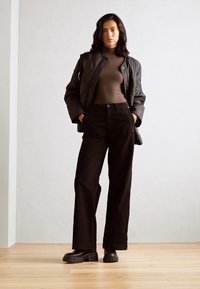 Padded črn plašč čez rjav rolak, v kombinaciji z visokim črnimi hlačami in močno črnimi škornji na lesenem tleh.