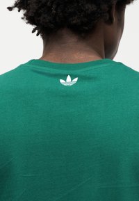 Πράσινο βαμβακερό μπλουζάκι με στρογγυλή λαιμόκοψη, με λευκό λογότυπο Adidas στο πίσω μέρος κοντά στη γραμμή του λαιμού. Ομαλή υφή, casual σχεδιασμός.