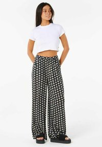 T-shirt bianco tagliato, pantaloni neri a gamba larga con motivo floreale bianco e sandali platform neri. Il tessuto appare morbido con un design fluido.