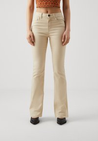 Mujer vestida con pantalones acampanados beige, zapatos de tacón puntiagudos negros y un top corto con patrón naranja, de pie contra un fondo liso.