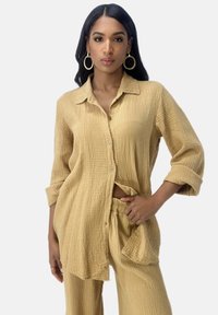 Camicia button-up beige chiaro realizzata in tessuto testurizzato con una vestibilità rilassata, abbinata a pantaloni larghi coordinati. Presenta maniche arrotolabili e tasche laterali.