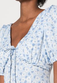 Světlé modré šaty s balónovými rukávy, květinový vzor v bílé a tmavší modré, výstřih do V a uvazovací detail v přední části. Texturovaná tkanina.