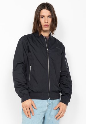 Personne aux cheveux bruns mi-longs portant une veste bomber noire zippée et un jean bleu clair, debout devant un fond blanc.