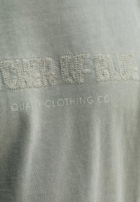 Close-up van lichtgrijze stof met geborduurde tekst "CHER OF BLUE" en daarop gedrukte tekst "QUALITY CLOTHING CO" eronder.