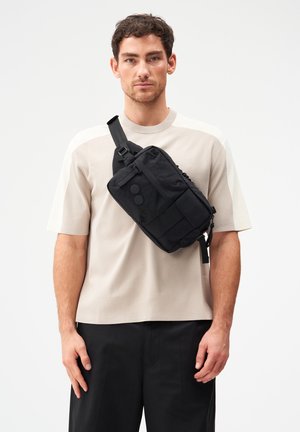 Mann trägt beige kurzärmeliges Hemd und schwarze Hose mit schwarzer Umhängetasche, die mehrere Taschen und zwei runde Knöpfe aufweist.