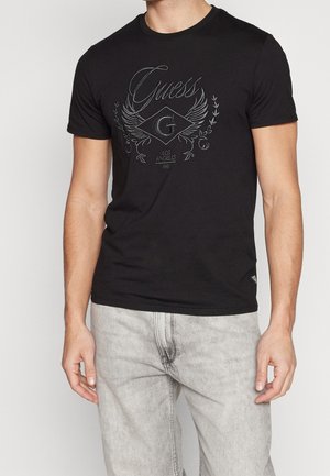 Homme portant un t-shirt noir Guess avec un logo ailé "G" et un texte "Los Angeles 1981", associé à un jean gris clair.