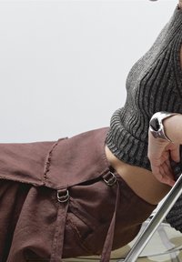 Personne portant une jupe marron avec des bretelles à boucle métallique et un haut gris côtelé, appuyée sur une chaise en métal, portant un grand bracelet argenté avec une pierre noire.