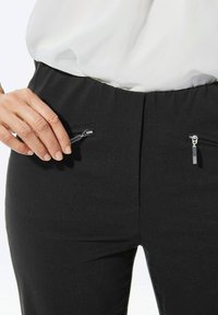 Un pantalon noir avec une texture lisse et deux poches zippées à l'avant. La taille est plate, offrant un aspect élégant.