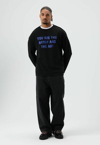Schwarzer Pullover mit blauem Text "DU BIST DER KÜNSTLER UND DIE KUNST", kombiniert mit lockeren schwarzen Jeans und klobigen schwarzen Schuhen mit pinken Akzenten.