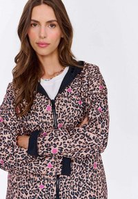 Žena s dlouhými hnědými vlasy má na sobě růžovou a černou leopardí zipovou bundu, bílou košili a barevný korálkový náhrdelník, ruce má založené v pase.