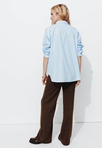 Chemise surdimensionnée bleu clair avec des manches retroussées, associée à un pantalon large marron pailleté et des chaussures noires. Design minimaliste, couleurs unies.
