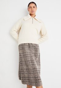 Kremfarget strikket genser med stor krage og halv-glidelås, kombinert med et midtlangt rutete skjørt i brune og beige toner. Strukturfabric.