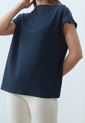 Mujer con una blusa azul marino de manga corta y suelta con detalles de botones dorados en los hombros y pantalones de color crema, de pie frente a un fondo liso.