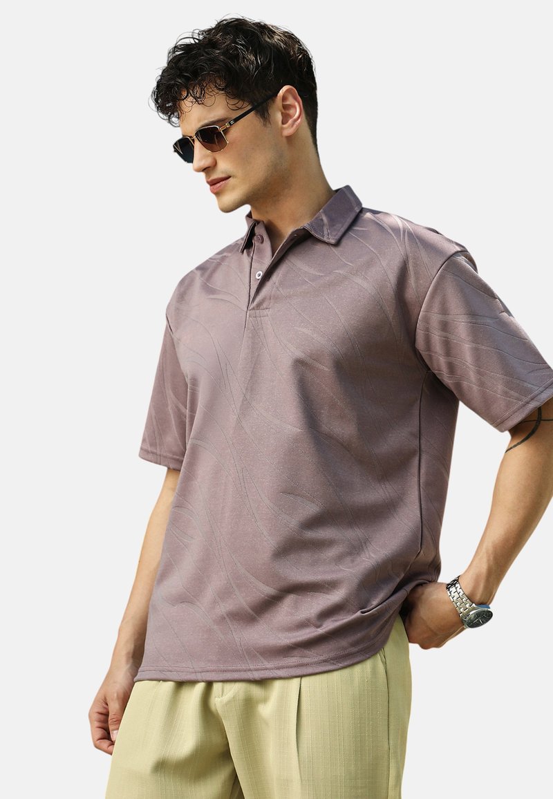 Koszula polo z krótkim rękawem w stonowanym kolorze mauve, z delikatnymi falistymi wzorami, kołnierzykiem i zapięciem na dwa guziki. Zestawiona z jasnymi spodniami.