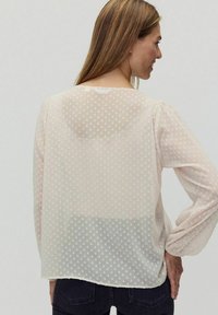 Gennemsigtig, lys beige bluse med strukturerede prikker, lange voluminøse ærmer og en rund halsudskæring. Bagsiden viser en let længere bund.