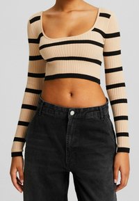 Kobieta w beżowym prążkowanym topie crop z czarnymi poziomymi paskami i czarnych spodni wysokiej talii, pokazana od ramion do bioder.