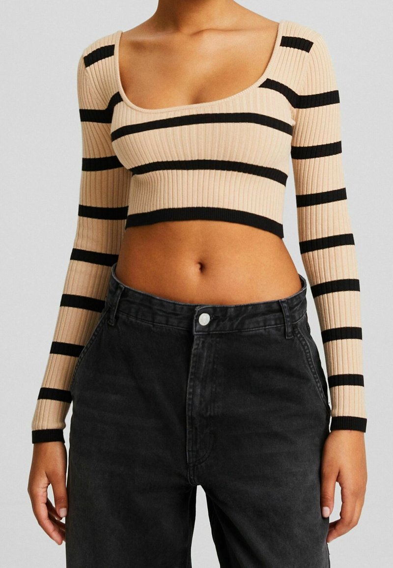 Kobieta w beżowym prążkowanym topie crop z czarnymi poziomymi paskami i czarnych spodni wysokiej talii, pokazana od ramion do bioder.