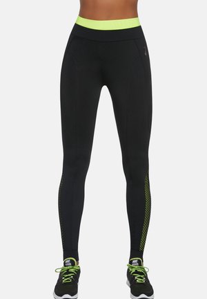 Osoba ubrana w czarne legginsy z neonowo zielonym pasem w talii i bocznymi akcentami, w połączeniu z czarnymi i zielonymi butami do biegania marki Nike, stojąca prosto.