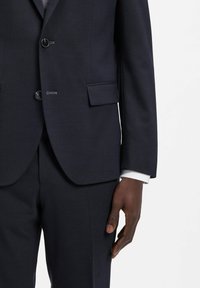 Abito blu navy con tessuto strutturato, chiusura a un bottone e tasca con pattina. Polsini della camicia bianca visibili sotto la manica della giacca.