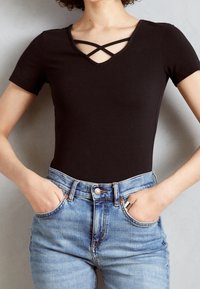 Svart kortärmad body med V-hals och korsade band, tillsammans med hög midja blå denimjeans, händerna i fickorna.