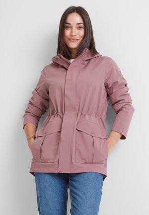 Woman wearing mauve hooded jacket with buttoned flap pockets and cinched waist, paired with blue jeans, standing against a white background.