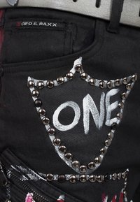I pantaloni in denim neri presentano un design bianco "ONE" bordato da borchie metalliche. Inclusa una tasca con etichetta del logo e un dettaglio con zip.