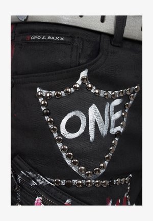 Schwarze Jeans mit einem weißen "ONE"-Design, umrandet von metallischen Nieten. Verfügt über eine Tasche mit einem Logo-Aufnäher und einem Reißverschlusdetail.