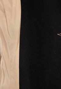 Tofarvet beklædningsgenstand med et glat beige stof på den ene side og et struktureret sort uldlignende materiale på den anden, med kontrasterende sømme.