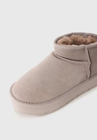 Papuci din suede bej deschis cu talpă groasă și căptușeală pufoasă vizibilă la deschiderea gleznei, pe un fundal alb.