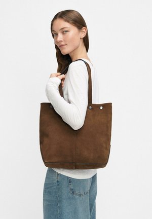 Frau in weißem Langarmshirt und blauen Jeans, die eine große braune Wildleder-Tasche über der Schulter trägt, vor einfarbigem Hintergrund.