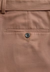 Gros plan sur une poche arrière marron de pantalon avec une fermeture à bouton noir et une boucle de ceinture visible au-dessus.