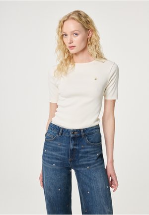 Blonde woman wearing white short-sleeve top and blue jeans with small heart patterns, standing against plain light background.