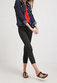 Giacca di jeans, blu scuro, design corto, con chiusura a bottoni, maniche arrotolate. Top rosso brillante, leggings neri aderenti e sandali infradito neri.