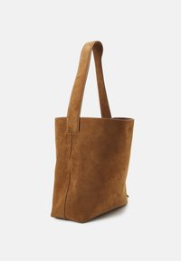 Sac cabas en daim marron avec deux anses, une large ouverture sur le dessus et une texture lisse. Présente des détails de coutures discrètes sur les côtés.