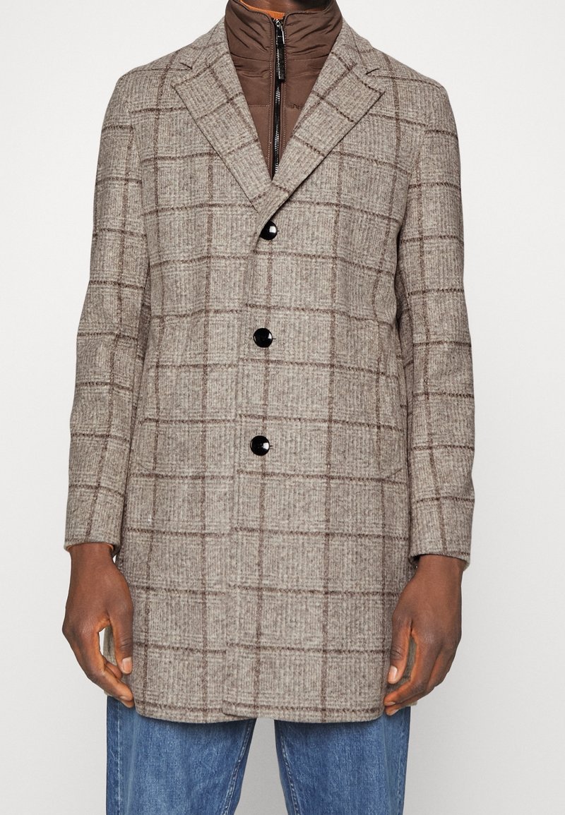 Homme portant un manteau en laine à carreaux beige par-dessus un pull zippé marron et un jean bleu, debout avec les mains détendues le long du corps.