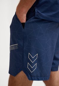 Calções atléticos na cor azul-marinho em tecido leve, apresentando um padrão geométrico com linhas roxas. Incluem bolsos laterais e um logo refletor.