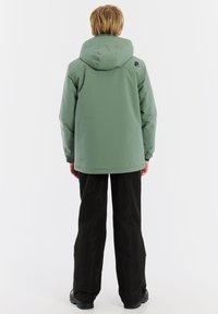 Grüne wasserdichte Jacke mit Kapuze, elastischen Bündchen und geradem Schnitt, kombiniert mit schwarzen Hosen und robusten schwarzen Schuhen.