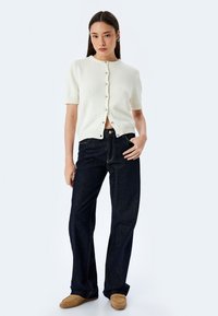 Cardigan bianco a maglia con maniche corte, scollo rotondo e chiusura frontale con bottoni, abbinato a jeans a gamba larga blu scuro e mocassini marroni.