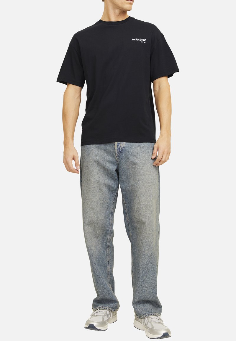 Maglietta nera in cotone, maniche corte, con la scritta "PARADISE". Indossata con jeans in denim azzurro chiaro, vestibilità comoda, senza fantasia.
