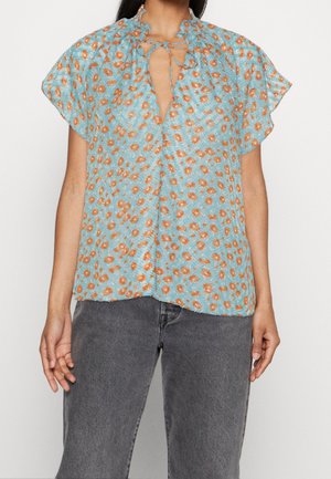 Femme portant un chemisier bleu clair à manches courtes avec un motif floral orange et un col à nouer, associé à un jean gris foncé.