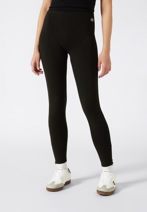 Persona con leggings negros de cintura alta con un pequeño logo de Champion cerca de la cintura, calcetines blancos de Champion y zapatillas blancas con suelas beige y marrones.