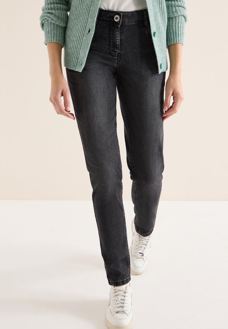 Osoba ubrana w wyblakłe czarne obcisłe dżinsy, białe trampki i miętowy zielony dziergany kardigan z białą koszulą pod spodem.