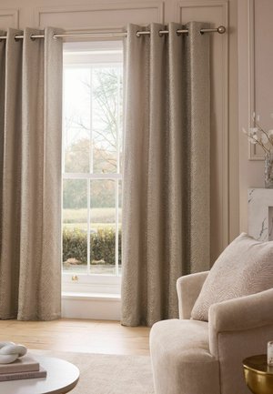 Rideaux beige à motifs suspendus à une tringle en métal à côté d'une fenêtre avec un fauteuil beige et un coussin assorti à proximité.