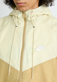 Light yellow and tan Nike windbreaker featuring a chevron design, zipper closure, adjustable hood with drawstrings, and embroidered logo.