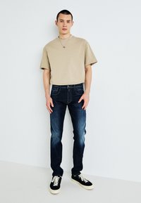 Beige t-paita lyhyillä hihoilla yhdistettynä tummansinisiin slim-fit farkkuihin ja mustiin lenkkareihin. Farkuissa on hienovarainen kulutus ja klassinen 5-taskuinen muotoilu.