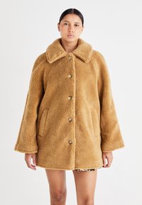 Rjav, puhast shearling plašč s širokim ovratnikom, ohlapnim krojem, sprednjimi žepi in gumbnim zapiranjem. Strukturirana površina in topel videz.