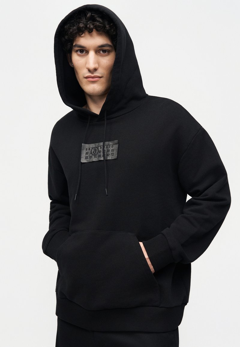 Junger Mann mit lockigem Haar, der einen schwarzen Hoodie mit Fronttasche und kleinem Brustaufnäher trägt, Hände in den Taschen, vor einem schlichten hellen Hintergrund.