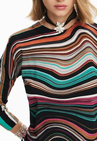 Femme portant un pull à rayures multicolores, un collier ras-du-cou floral argenté sur ruban noir, et un bracelet manchette argenté au poignet gauche.