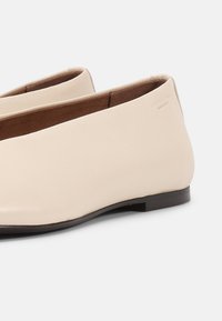 Δερμάτινα παπούτσια χωρίς κορδόνια σε κρεμ απόχρωση με στρογγυλεμένες μύτες και μαύρες επίπεδες σόλες, εμφανιζόμενα από πλάγια γωνία σε απλό φόντο.